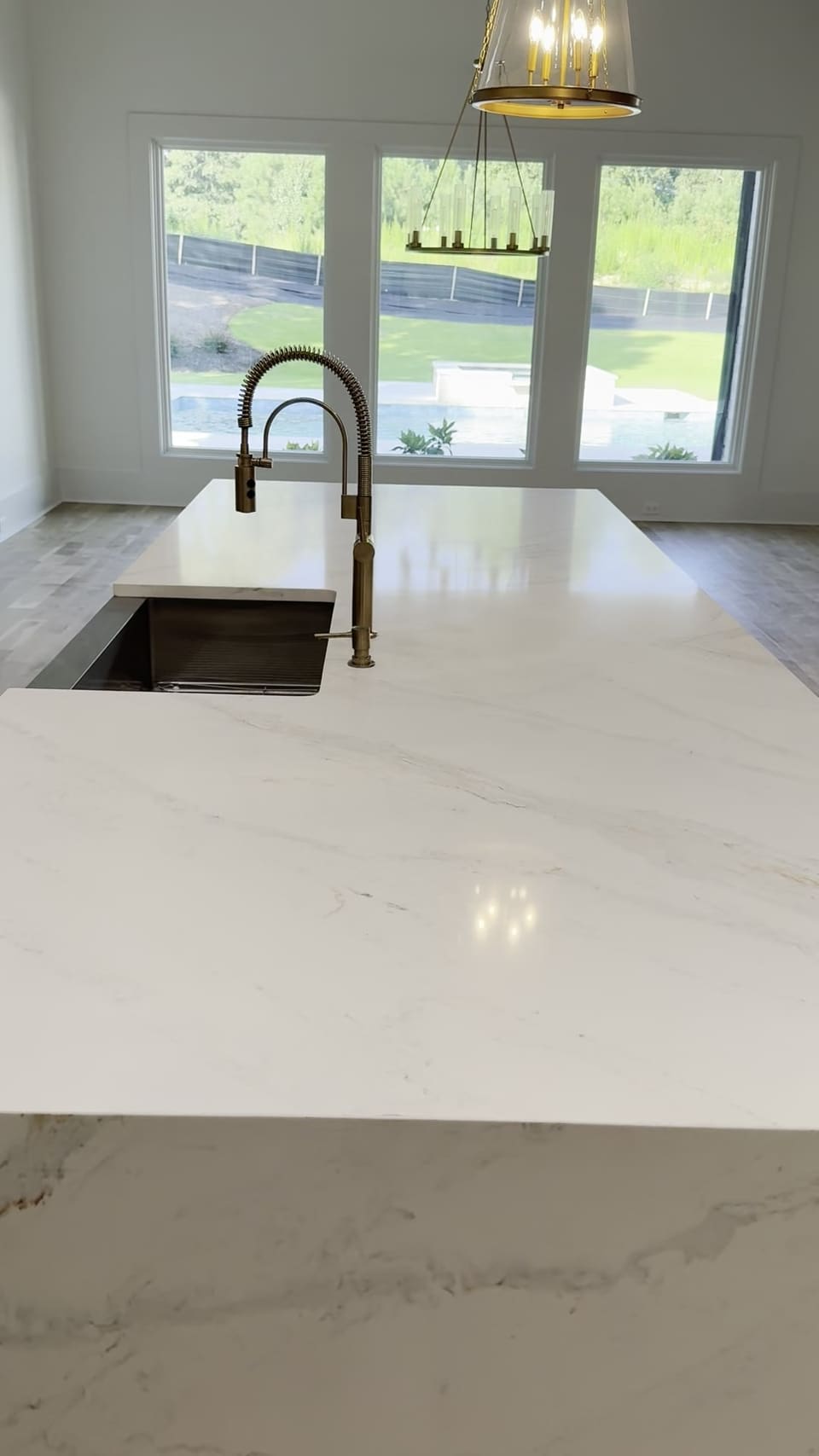 White stone island with a farmhouse sink and brass faucet facing a wall of windows