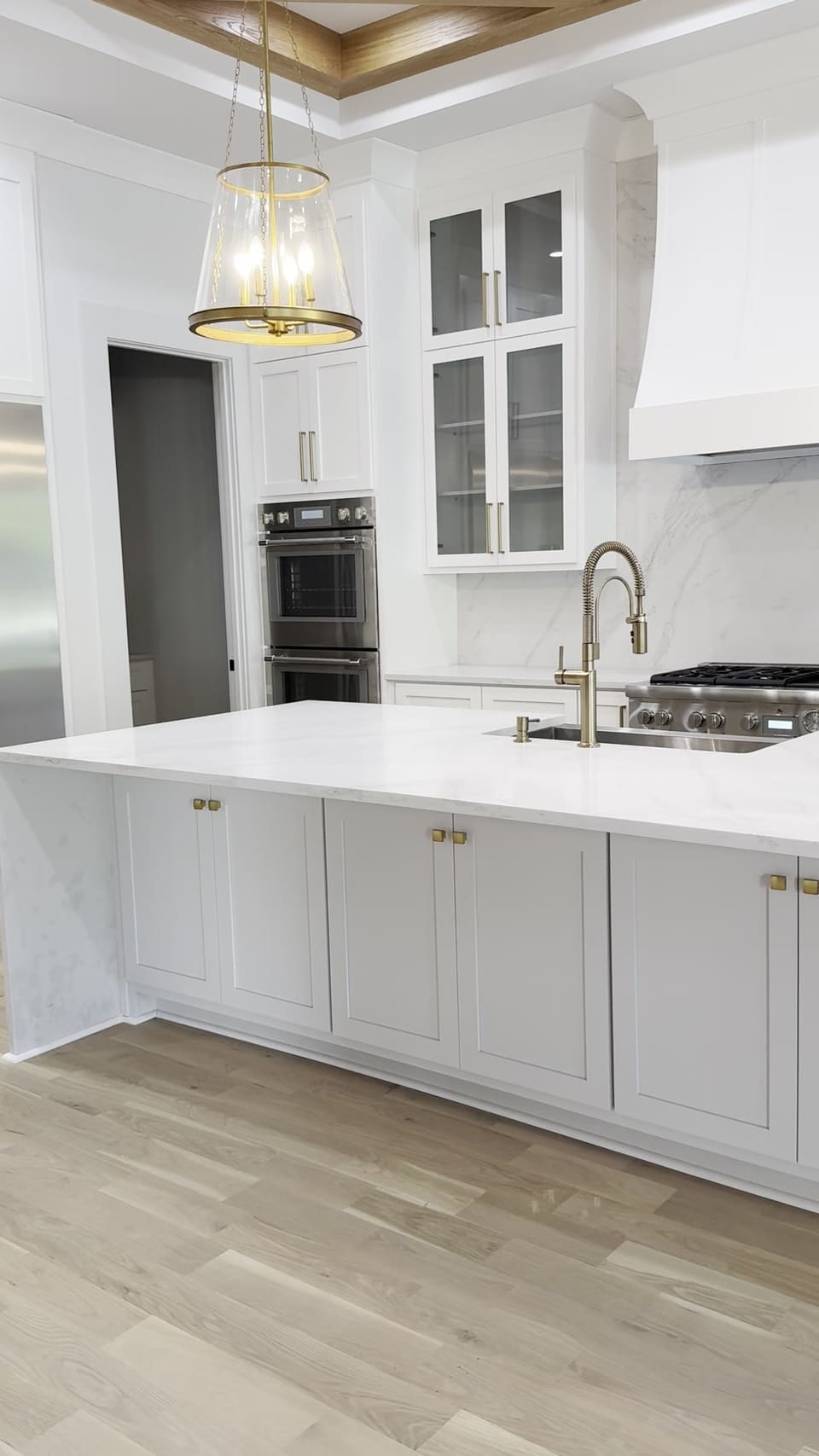 White island with a brass faucet in front of a slab backsplash and glass front cabinets under pendant lights