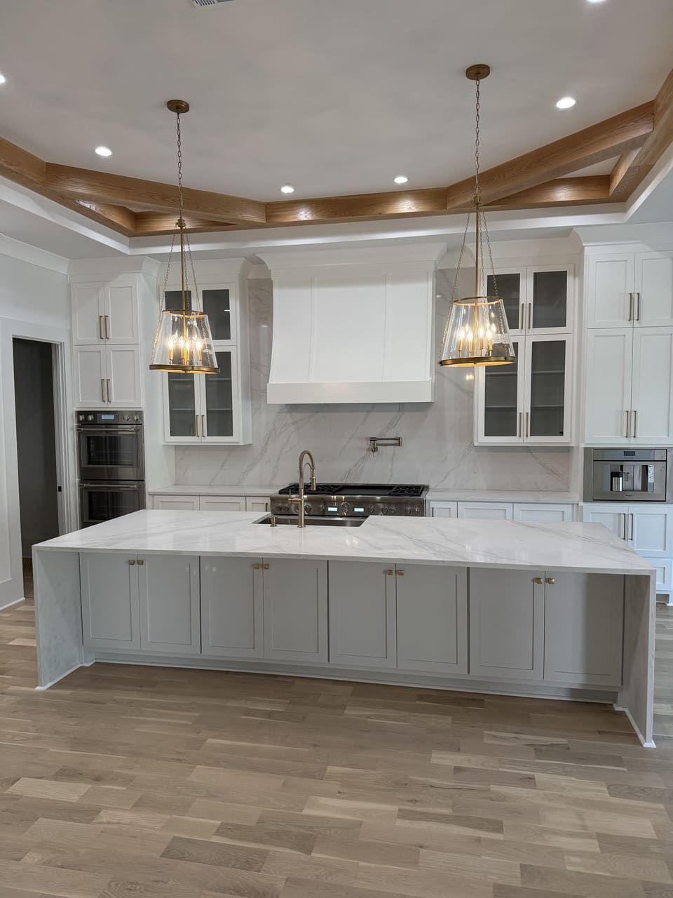 Long white waterfall island under glass pendants in a white kitchen with a coffered wood ceiling