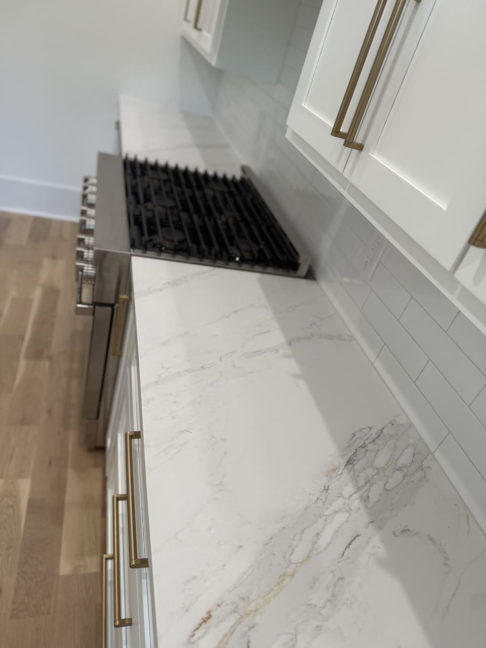Close view of a white veined countertop running beside a stainless gas rangetop and brass cabinet pulls