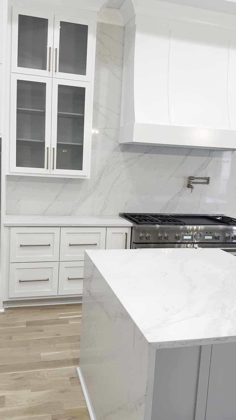 White waterfall island in front of a full slab backsplash, range and glass front cabinets